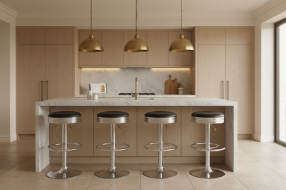 A modern kitchen island with a white marble countertop and four chrome bar stools with black seats, set in a bright kitchen with light wood cabinets and three gold pendant lights.