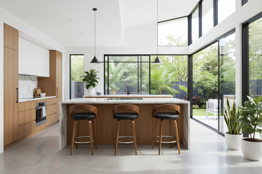 A modern kitchen island with a light wood grain cabinet base, a white marble countertop, and three wooden bar stools with black seat cushions, set in a bright contemporary kitchen with large windows and plants.