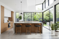 A modern kitchen island with a light wood grain cabinet base, a white marble countertop, and three wooden bar stools with black seat cushions, set in a bright contemporary kitchen with large windows and plants.
