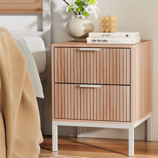 Wooden bedside table with two drawers, books, and decorative items next to a bed.