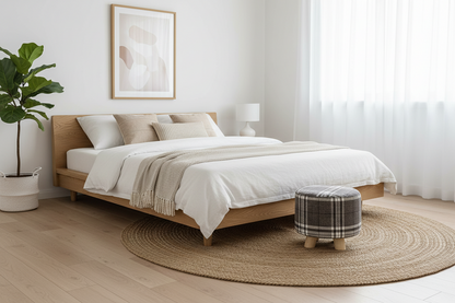 A modern light wood platform bed dressed in white linens and a beige throw blanket, styled in a bright minimalist bedroom with a round woven jute rug, a plaid ottoman, a potted plant, and a window with sheer curtains.