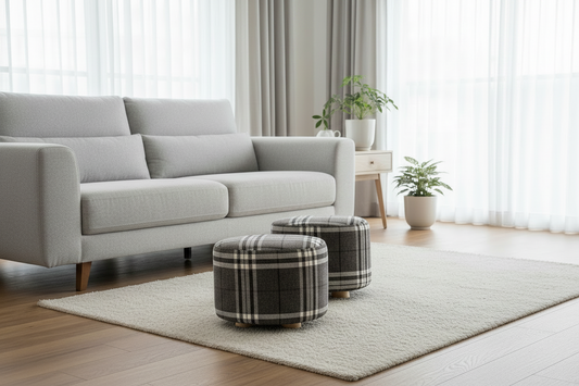 A modern living room scene with a light gray sofa, two plaid ottomans on a cream rug, wood flooring, sheer curtains, and potted plants.