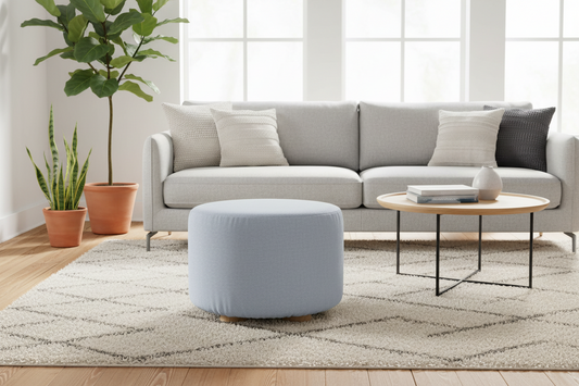 A modern living room scene featuring a light blue cylindrical ottoman in the foreground, set on a patterned beige rug in front of a light gray sofa. A round wooden coffee table with black metal legs sits to the right, and potted plants are visible on the left side of the room.