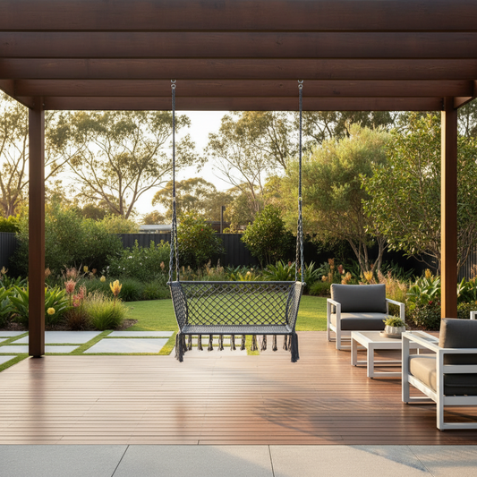 A dark gray outdoor porch swing hanging under a wooden pergola on a tiled patio, with matching patio armchairs and tables and a lush green garden in the background.