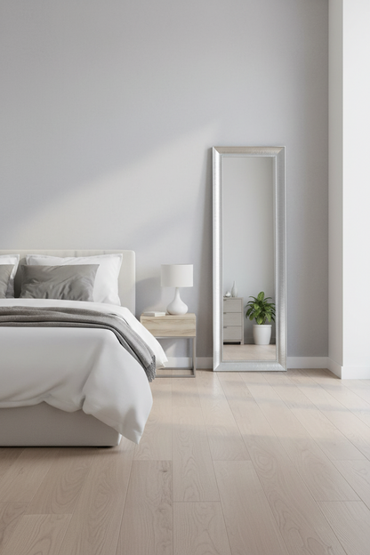 A tall, full-length floor mirror with a slim silver frame stands in a bright, modern bedroom next to a bed with white bedding and a small bedside table.