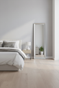 A tall, full-length floor mirror with a slim silver frame stands in a bright, modern bedroom next to a bed with white bedding and a small bedside table.