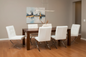 A modern dining room setup featuring a dark wood rectangular table surrounded by six white leather chairs with chrome legs, set against a warm gray wall with a framed abstract painting.