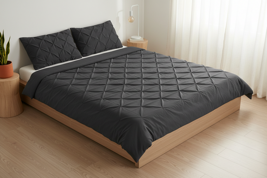 A bed dressed in a dark charcoal gray quilted duvet cover and matching pillowcases, set in a modern bedroom with light wood flooring, a wooden bed frame, and sheer white curtains.