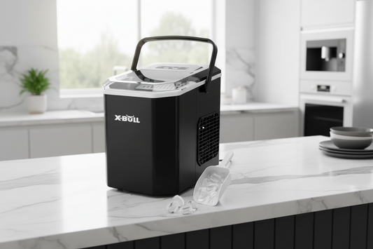 A black X-Bull portable ice maker with a clear lid and handle sits on a white marble kitchen countertop, with ice cubes and a clear scoop in front of it.