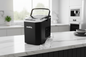 A black X-Bull portable ice maker with a clear lid and handle sits on a white marble kitchen countertop, with ice cubes and a clear scoop in front of it.